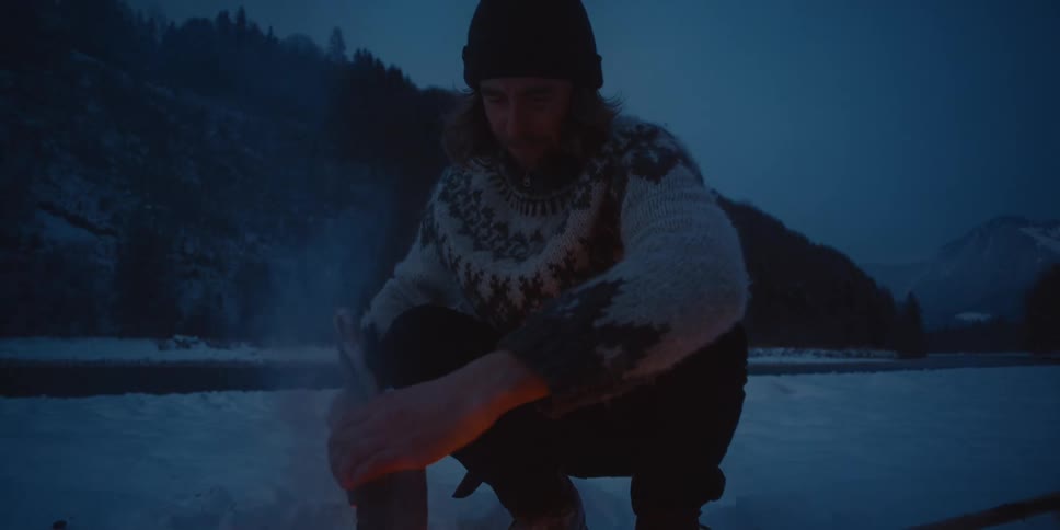 A man is holding an ax as he bends down and builds a campfire with pieces of split wood outside on a winter evening