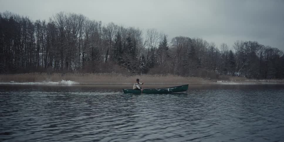 A man is rowing a canoe across the water and there is a forest of bare trees near the shore
