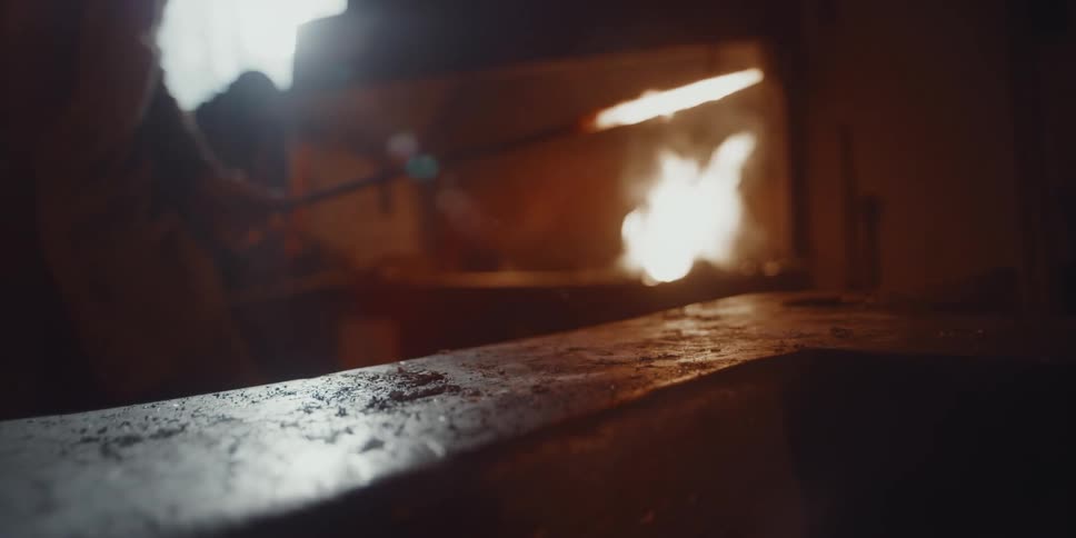 A man takes a burning hot metal object from a roaring forge and hits it with a hammer to give it shape
