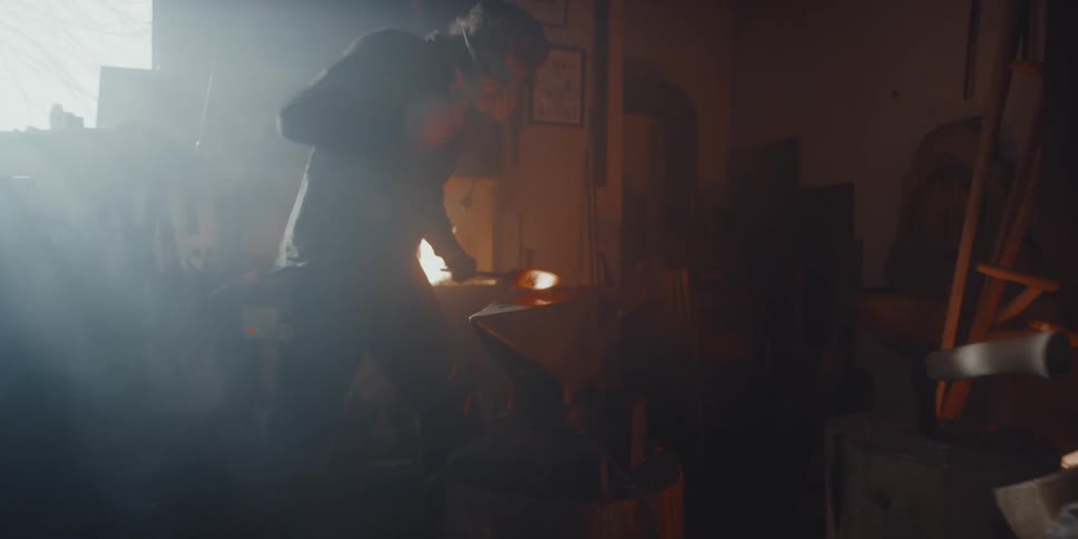 A man uses a hammer to hit a piece of flaming metal hard over an anvil