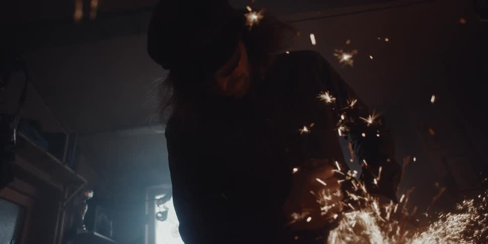 A man is working in a metal shop and sparks are glowing as they fly in the darkness