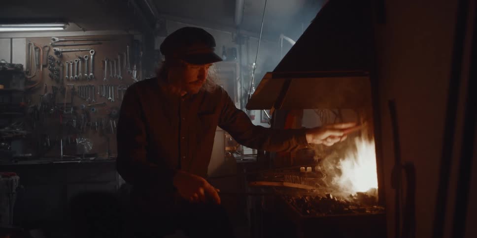A man in his workshop with tools hanging on the wall tending to a fire