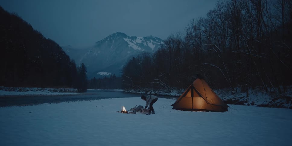 A man carries firewood to a campfire near a tent along a frozen river surrounded by bare trees and mountains in the distance