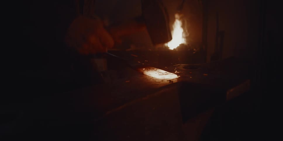 The flames in a fireplace burn in the darkness as a blacksmith hammers a piece of molten hot metal
