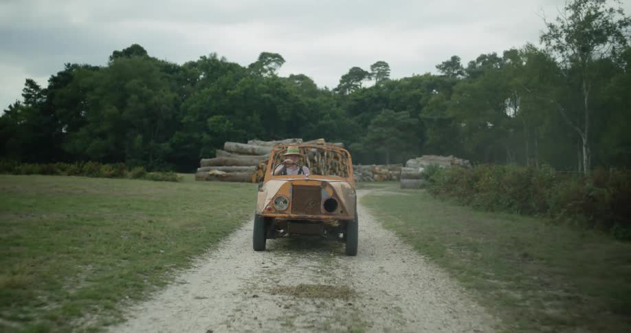 A man wearing a sunhat is driving a small orange vehicle on a dirt road in the country and there is a pile of logs in the distance