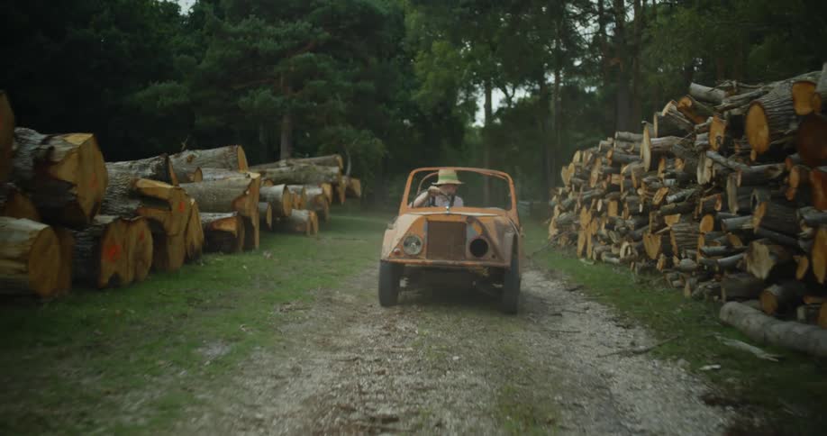 A man wearing a sunhat is driving a small orange vehicle on a dirt road surrounded by logs stacked high along the roadsides and there are trees in the distance