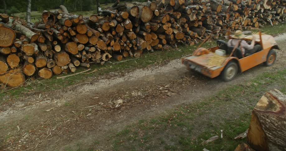 A man wearing a sunhat is driving a small orange vehicle on a dirt road by tall stacks of logs and firewood