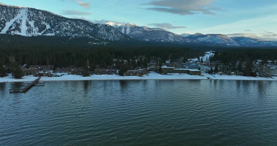 Snow covers the ground around buildings along the river valley as traffic moves on the streets and there are forest and mountains in the distance