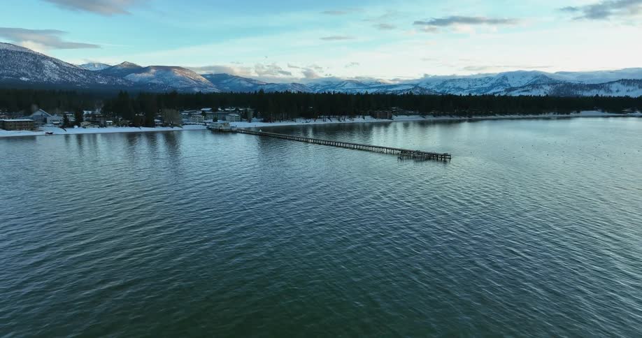 A pier stretches across the lake as the sky and trees reflect on the rippling water and there are mountains in the distance