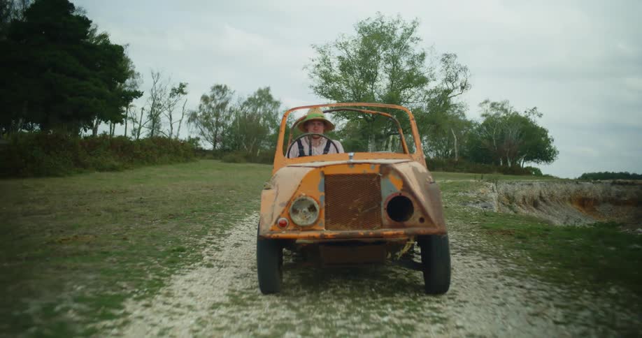 A farmer wearing a straw hat drives a small orange vehicle on a dirt road in the country