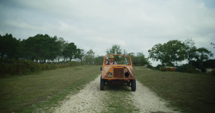 Man drives jeep with missing headlight along dirt road past a herd of cattle on a cloudy day