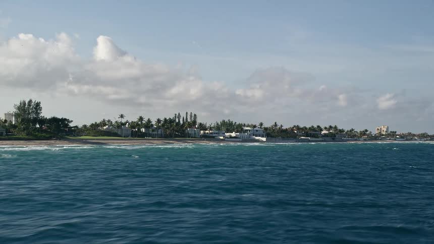 Aerial footage of homes along the shoreline of Palm Beach Florida with a mix of smaller low-rise buildings and taller structures visible in the distance and partly cloudy skies above