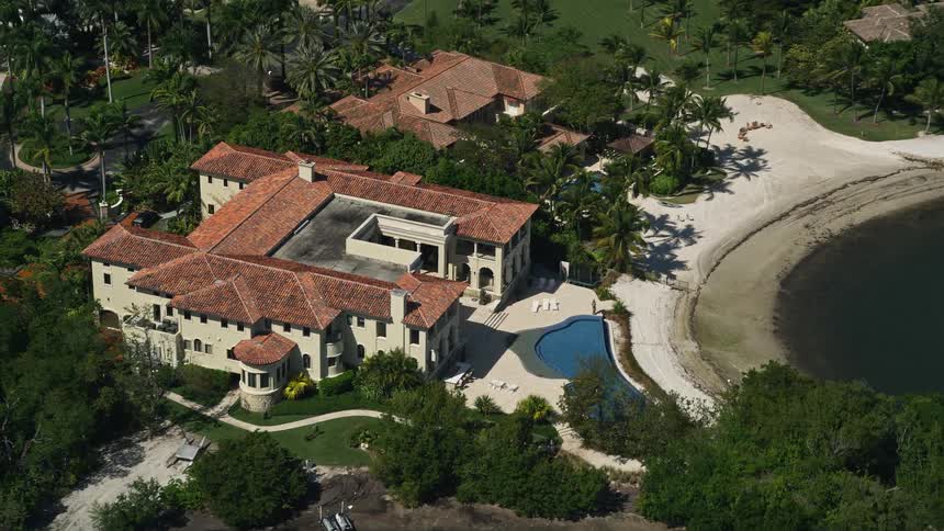 Aerial footage of a luxurious waterfront mansion with a pool surrounded by trees and a smaller neighboring mansion in Coral Gables Florida