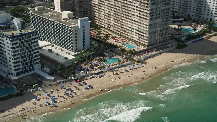 A stunning aerial view of sunbathers on the beach in front of Ocean Sky Hotel and Resort in Fort Lauderdale Florida with hotel buildings and pools in the background as waves crash against the coast