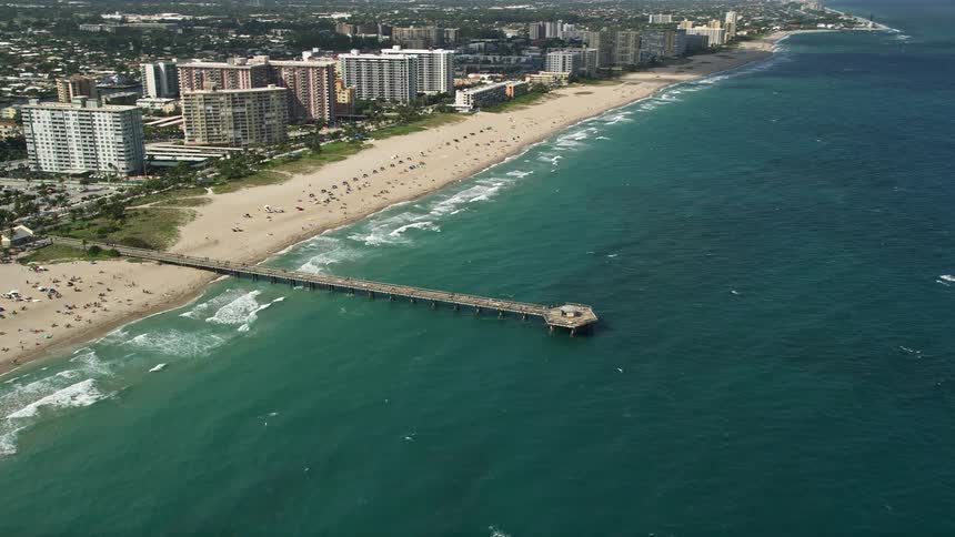 Approaching and orbiting Pompano Beach Pier capturing the lively recreational activities of sunbathers and beachgoers on a partly cloudy day in Pompano Beach Florida