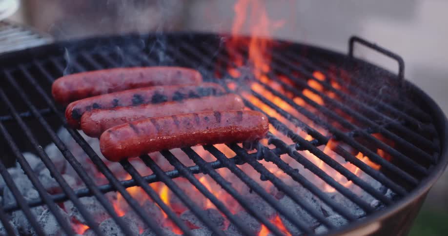 Four wieners are cooking in the flames of an outdoor grill in the daytime