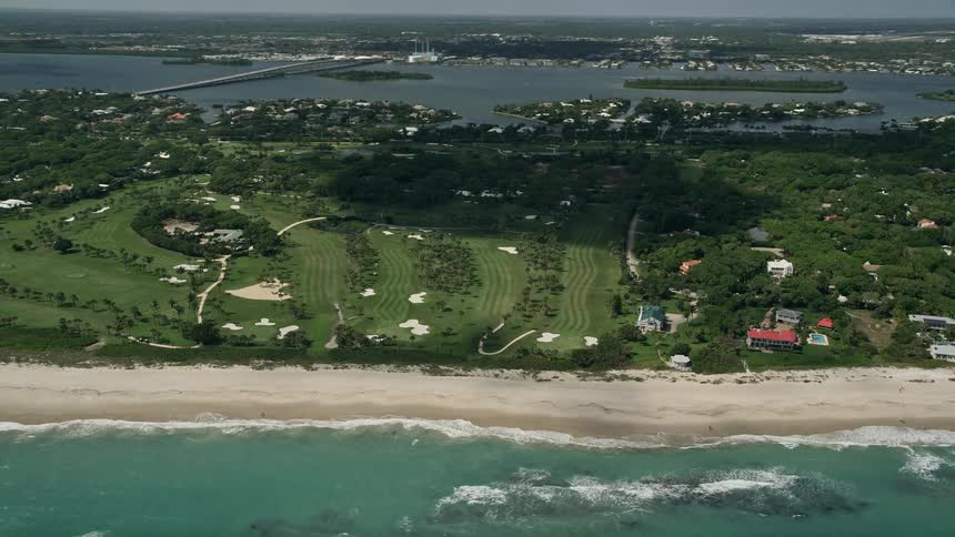 An aerial view of a partly cloudy coastline with a golf course flying away from the shore in Vero Beach Florida during summertime