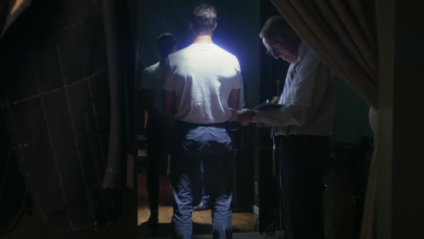 Man faces his reflection while standing under a spotlight as an older man reads from a book