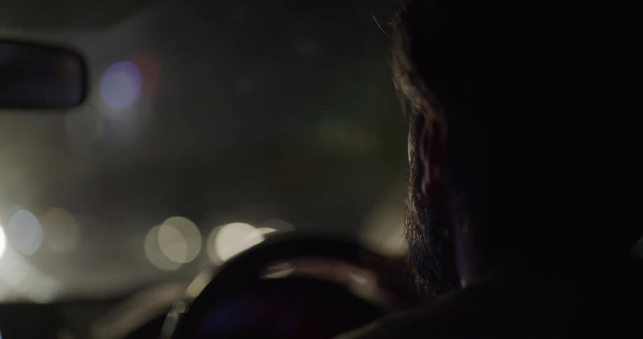 The side profile of a mans bearded face as he drives a vehicle at night as light flares shine through the window