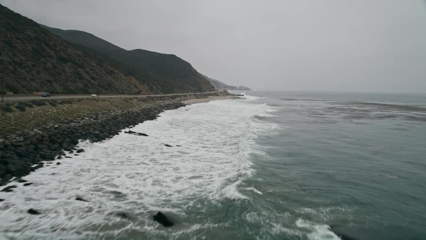 Low altitude aerial footage of waves crashing against the Malibu coast and Pacific Coast Highway on a hazy day