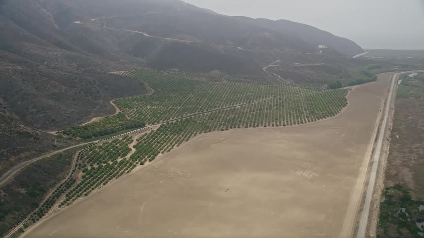 Aerial footage follows a rural road through light fog to approach crops and farmland in Point Mugu California