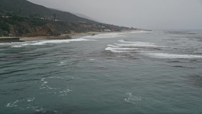A low altitude aerial footage showing waves crashing against coastal cliffs as the camera flies over the ocean towards a beach in Malibu California