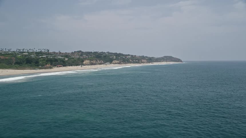 Aerial view of Point Dume in Malibu CA as the camera flies over blue waters near the beach on a hazy day