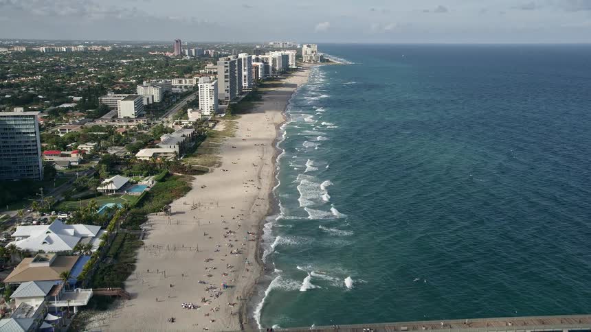 A stunning aerial footage of Deerfield Beach Florida showing a bustling beach with people and waves crashing and a pier extending out into the water