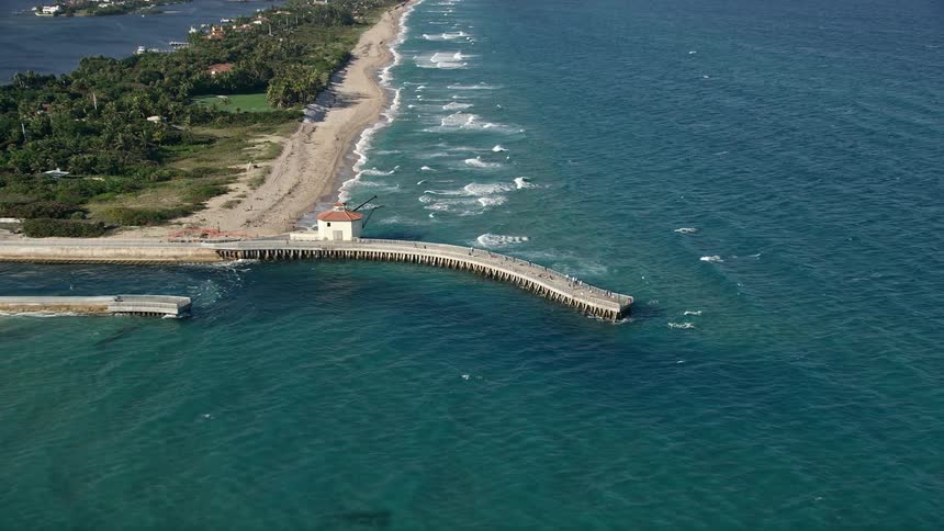 Approaching Boynton Beach Inlet by aerial footage tilting down to reveal a pier amidst a partly cloudy backdrop showcasing the recreational activities of beachgoers and sunbathers enjoying leisure activities in South Florida