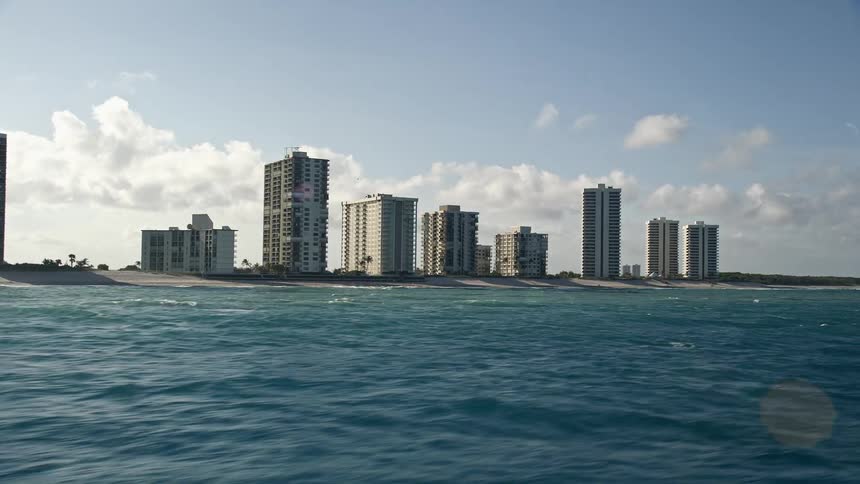 Flying over coastal condo complexes in Riviera Beach FL on a sunny day with a partly cloudy sky capturing the beautiful shoreline and oceanfront properties