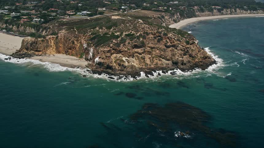 An aerial view captures the stunning California coastline by tilting from the ocean to reveal and approach the rugged cliffs of Point Dume in Malibu with sunshine and clear blue skies creating a scenic atmosphere perfect to fly over and capture the seasho
