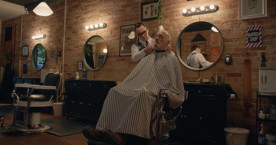 A barber trimming an elderly mans hair sitting in the chair while the man has his eyes closed