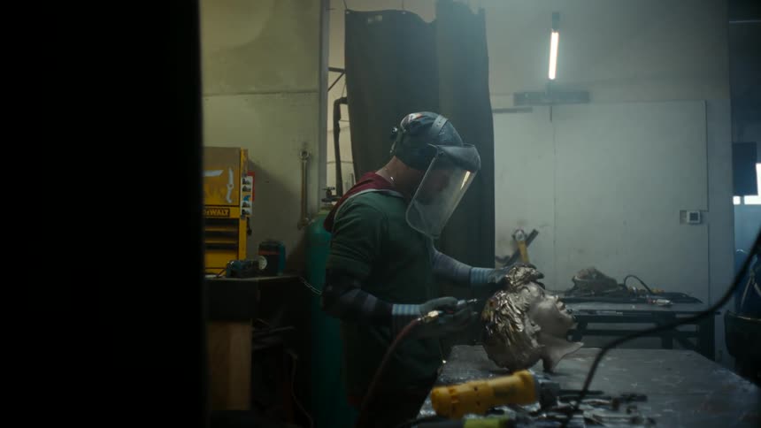A metal sculptor uses a drill to finetune detail on a metallic head sculpture