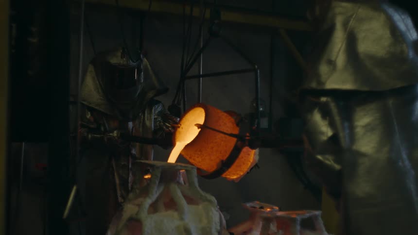 Two metalworkers wearing protective gear pour molten metal into molds