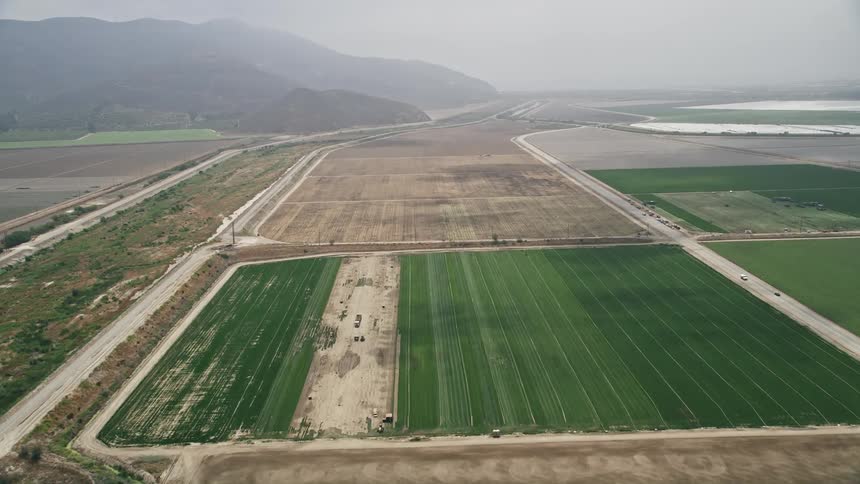 An aerial view fly over of farmland between country roads with light fog in the distance during a sunny day in Camarillo California