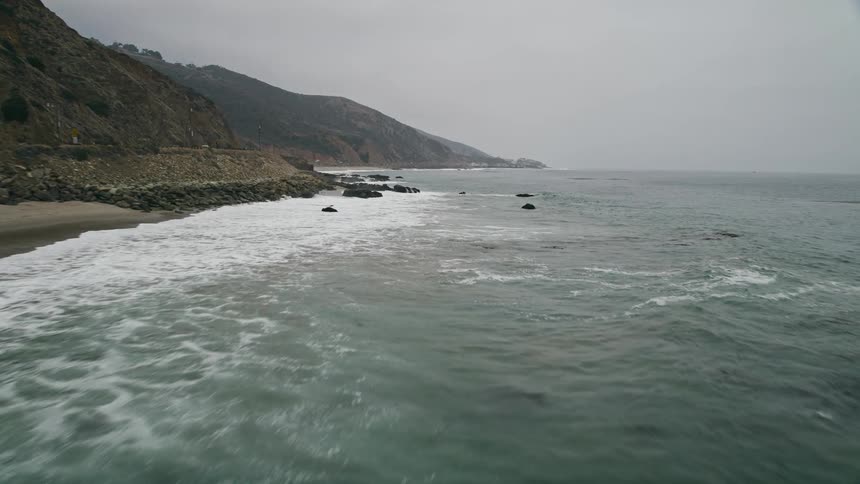 A stunning aerial view of waves crashing against the rocky coastline of Malibu California with kelp and haze in the foreground