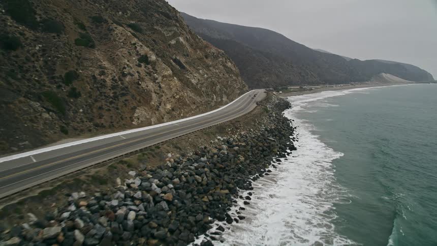 Aerial view of vehicles driving along Highway 1 near Point Mugu California hugging the mountains that border the coast with light traffic