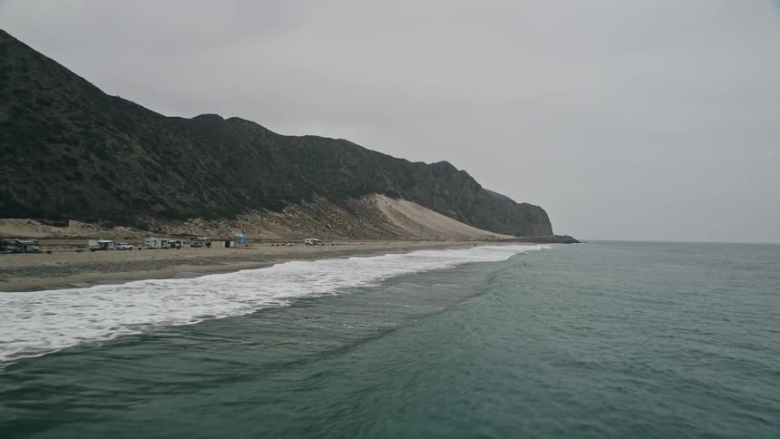 A low flying aerial clip of Pacific Ocean waves approaching a beach in Malibu California with a backdrop of mountains perfect for sightseeing beachgoers and leisure activities in Southern California