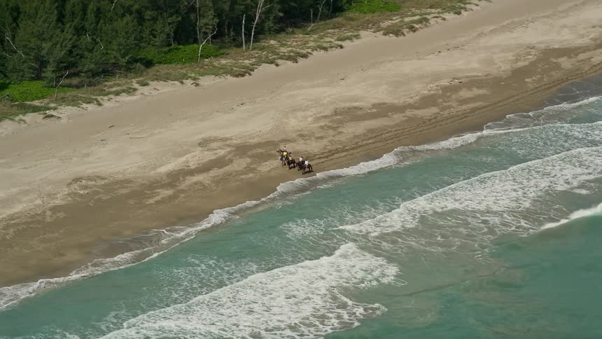 Horseback riders covered in shadows ride along a populated Florida beach in the daytime as the aerial footage captures the shore from afar and then starts orbiting away