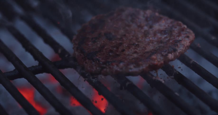 Smoke rises from glowing coals and a hamburger is flipped on a grill