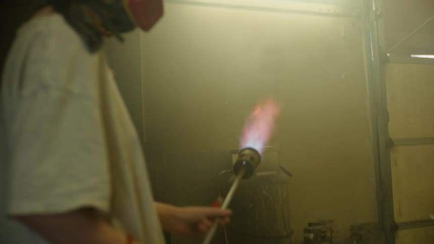 A man wearing protective head and face covering is standing in a room as he lights a blowtorch and a wild flame emerges
