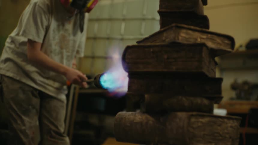 A person works on a metal piece of art work resembling a stack of books by using a blowtorch on it