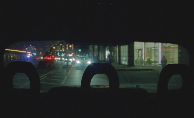 View of cars pedestrians storefronts city lights and buildings from a rear window of a vehicle as it is being driven on a city street at night