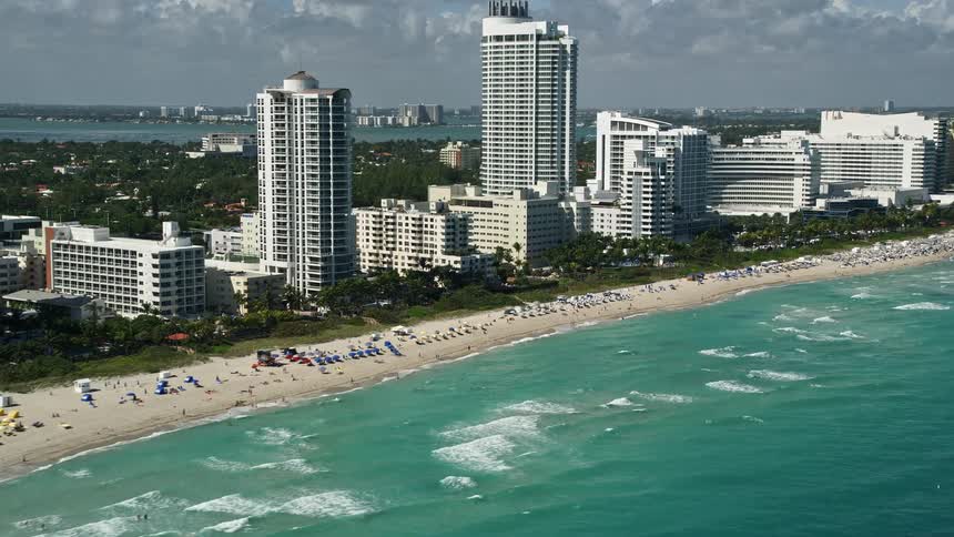 A bright and sunny day attracts beachgoers to Fontainebleau Miami Beach Florida with aerial footage showcasing nearby hotels and the bustling coastline