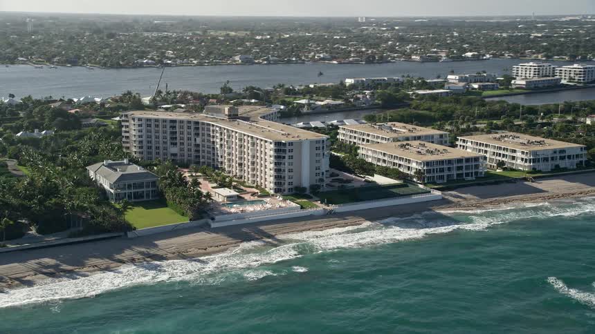 Birds eye view of a Palm Beach condominium complex located along the beach with waves crashing against the shoreline and clear skies in the background