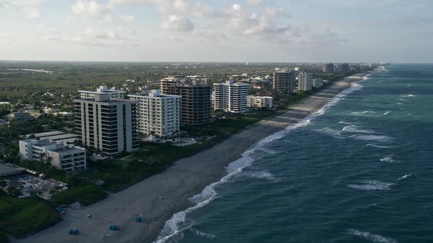 Aerial footage of Juno Beach Florida capturing condominium complexes casting a shadow over the beach while waves crash in