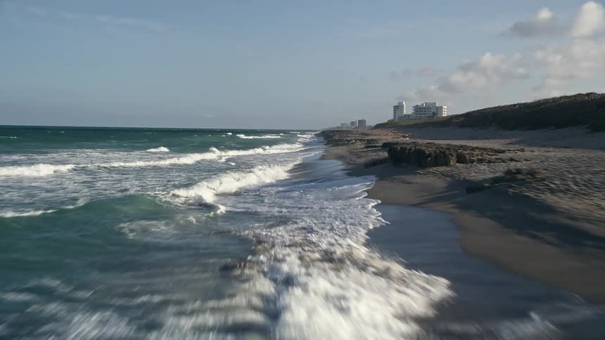 A low-flying aerial view of waves rolling onto an empty beach in Hobe Sound Florida during a partly cloudy day