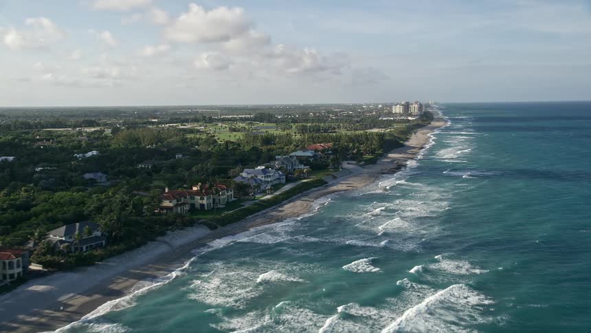 Aerial footage of North Palm Beach coastline passing by luxurious mansions and a popular beach showcasing the recreational activities of beachgoers during a partly cloudy afternoon