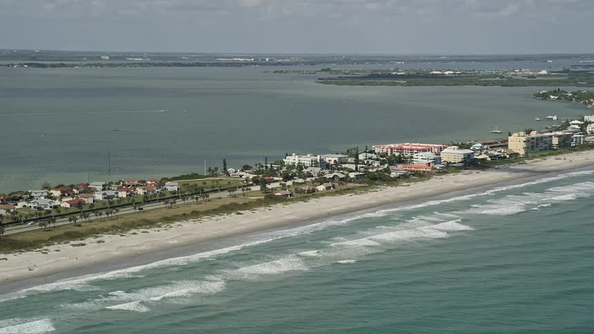 Aerial footage of a beach in Cocoa Beach Florida with waves crashing onto the shore and vehicles driving on the road parallel to the coast