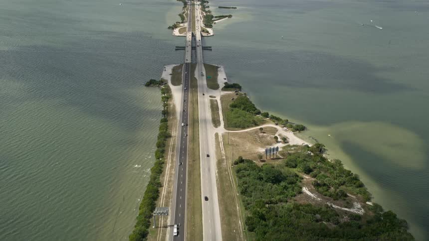 Aerial view of cars driving on a four-lane highway approaching Bennett Causeway revealing Merritt Island with natural rivers and waterways under partly cloudy skies on the east coast of Florida captured during spring or summer